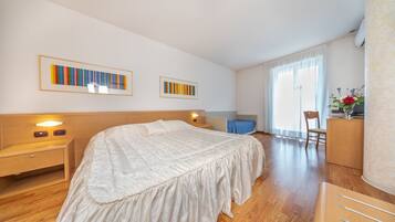 Ein Hotelzimmer mit einem großen Bett, einem Holz-Kopfteil, Nachttischen, einem Schreibtisch und einem Sessel. An der Wand befinden sich zwei gerahmte Kunstwerke, ein Fenster mit Vorhängen und ein kleiner Tisch mit einer Blumenvase.