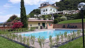 Una zona de piscina con un edificio identificado como 'Karinthall Camera Zimmer' al fondo, rodeado de vegetación y un cielo despejado.