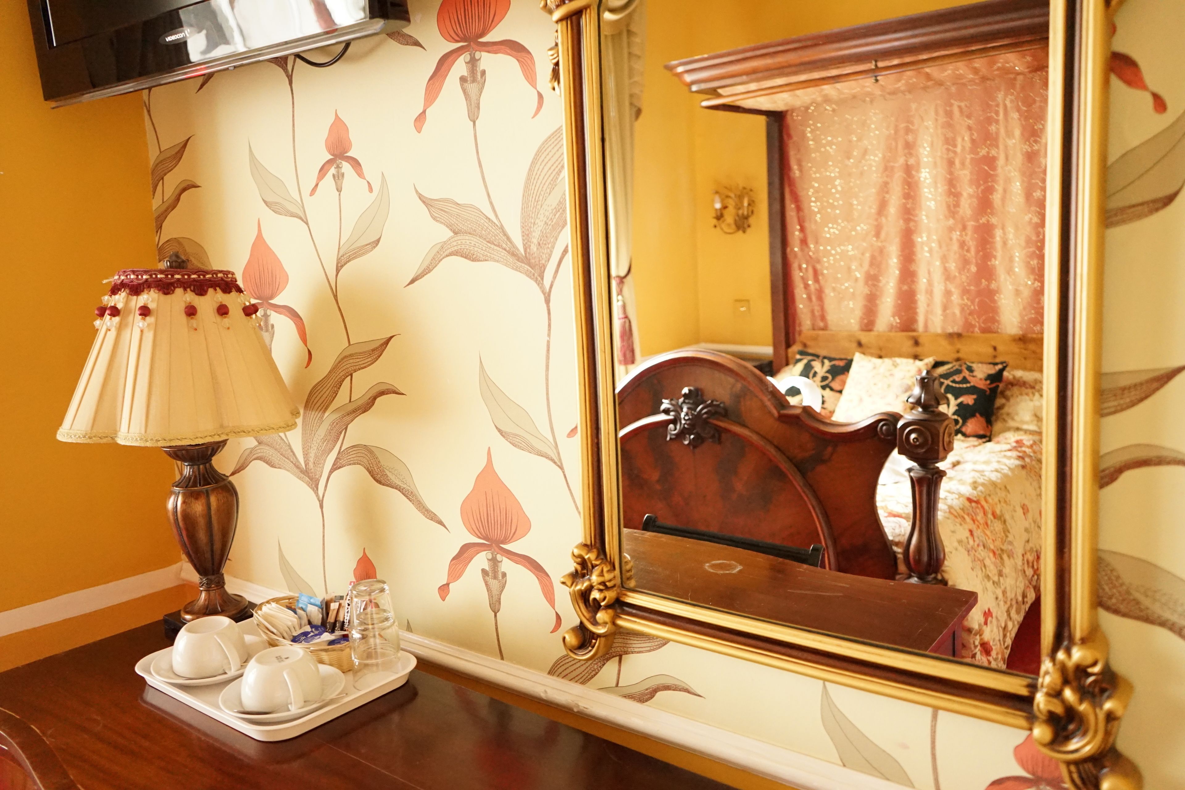 A hotel room with a wooden bed, a mirror, a tray with cups and a teapot, and a wall with floral wallpaper.