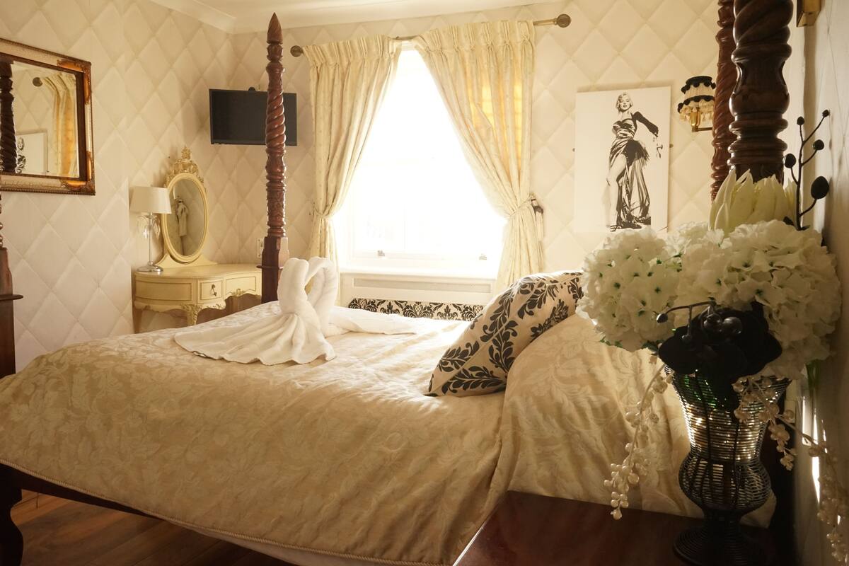 A bedroom with a four-poster bed, a bedside table with a lamp, a mirror, and a vase with flowers.
