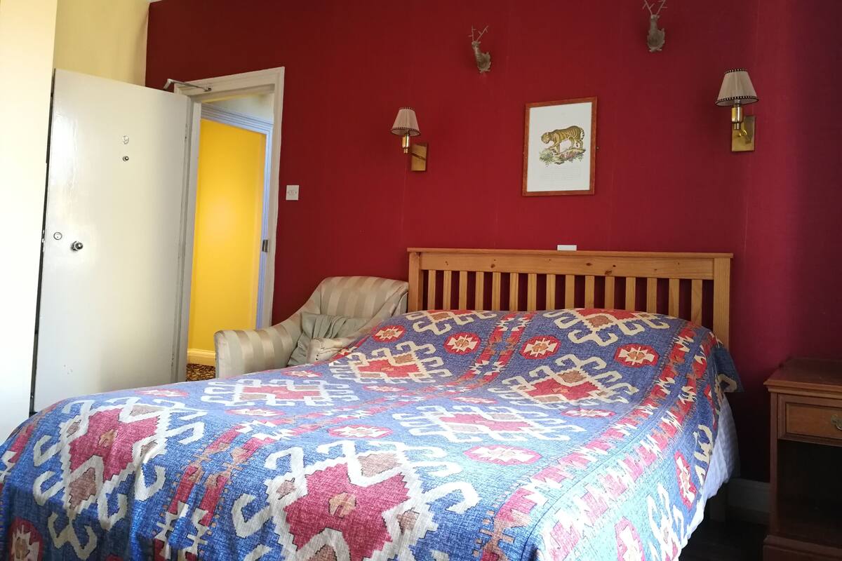 A bedroom with a wooden bed, a patterned blanket, a red wall, and two wall-mounted lamps.