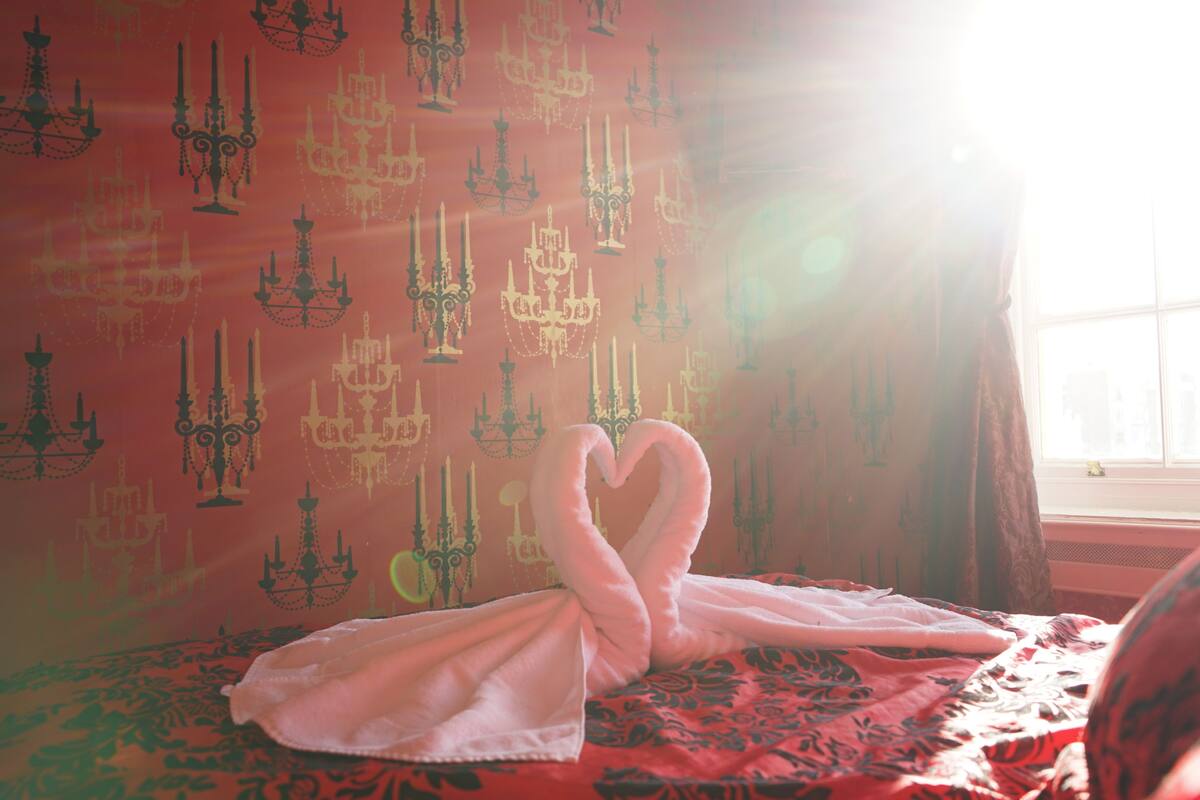 A room with a patterned wallpaper, a bed with a heart-shaped towel, and a window with sunlight.