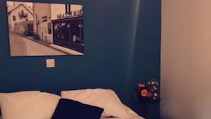 A double bed with a beige bedspread and two folded white towels. There is a dark blue pillow and a small table with a vase of flowers. A black and white photograph is mounted on the wall above the bed. - The New Inn (Beverley)