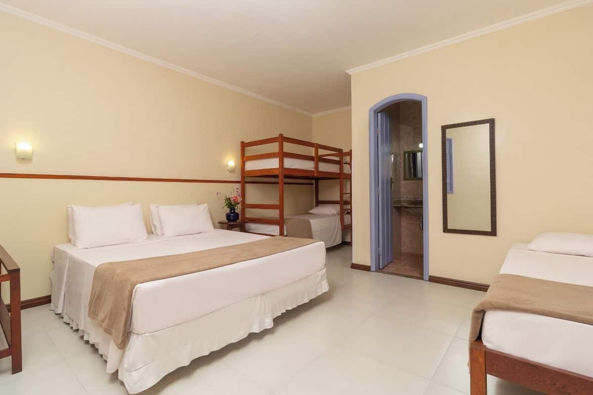 A hotel room with a bunk bed, a single bed, a bathroom with a mirror, and a small table with a vase.