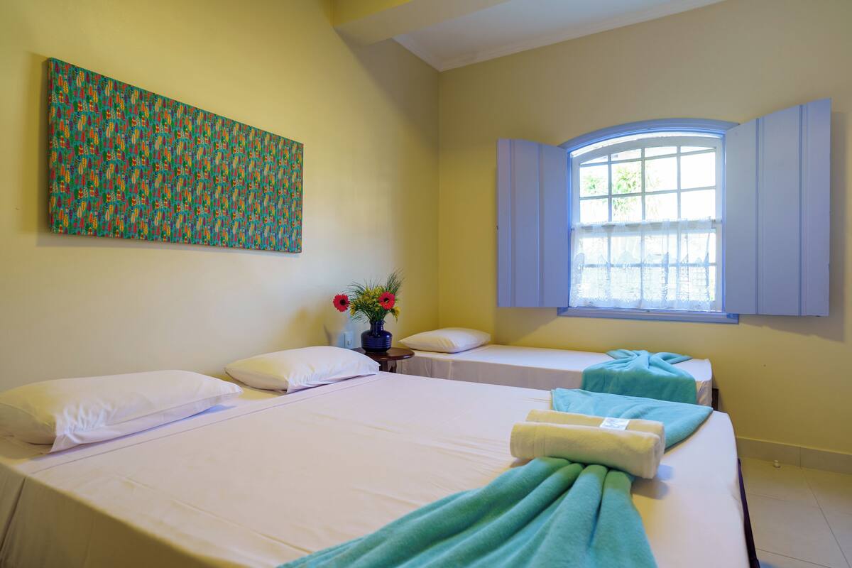 A hotel room with two beds, a large abstract painting on the wall, a window with curtains, and a vase with flowers on a nightstand.