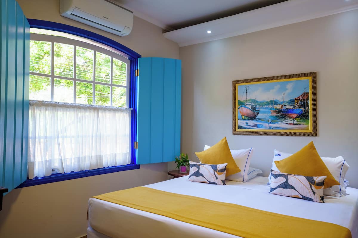 A hotel room with a bed, a window with blue curtains, a painting of boats, and a potted plant.