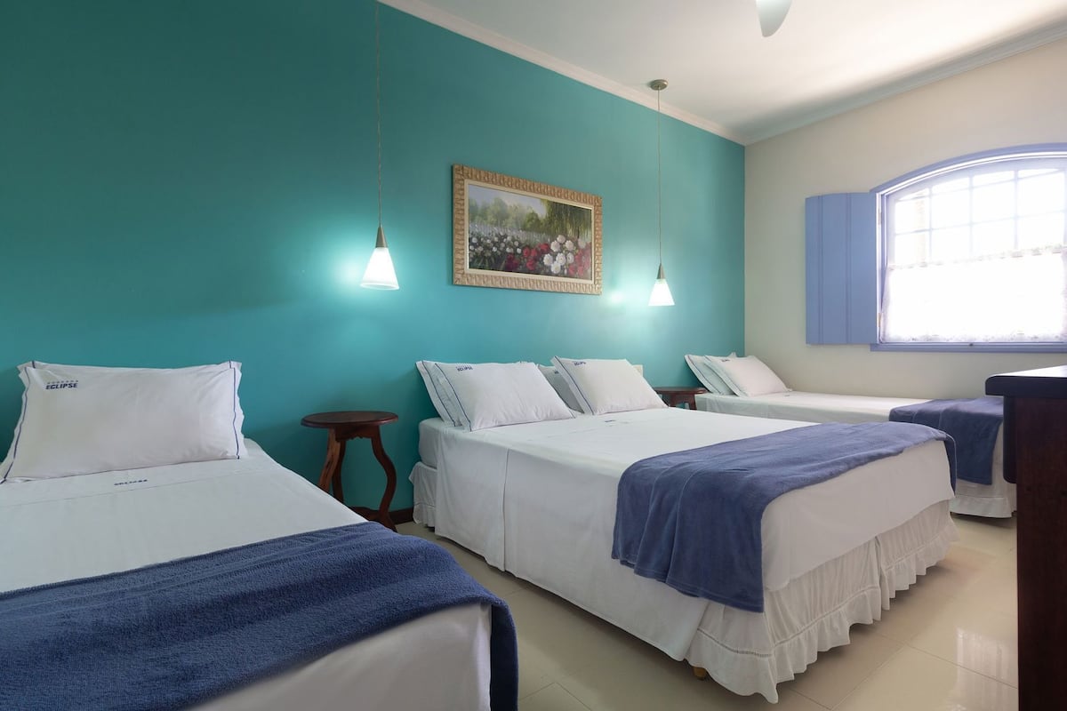 A hotel room with three beds, a small round table, a painting on the wall, and a window with blinds.