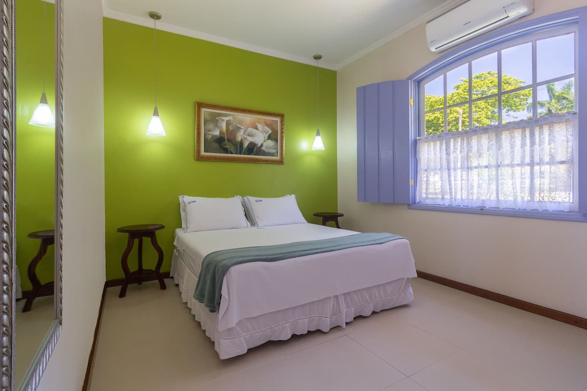 A bedroom with a bed, a window with lace curtains, a green wall, and a painting.