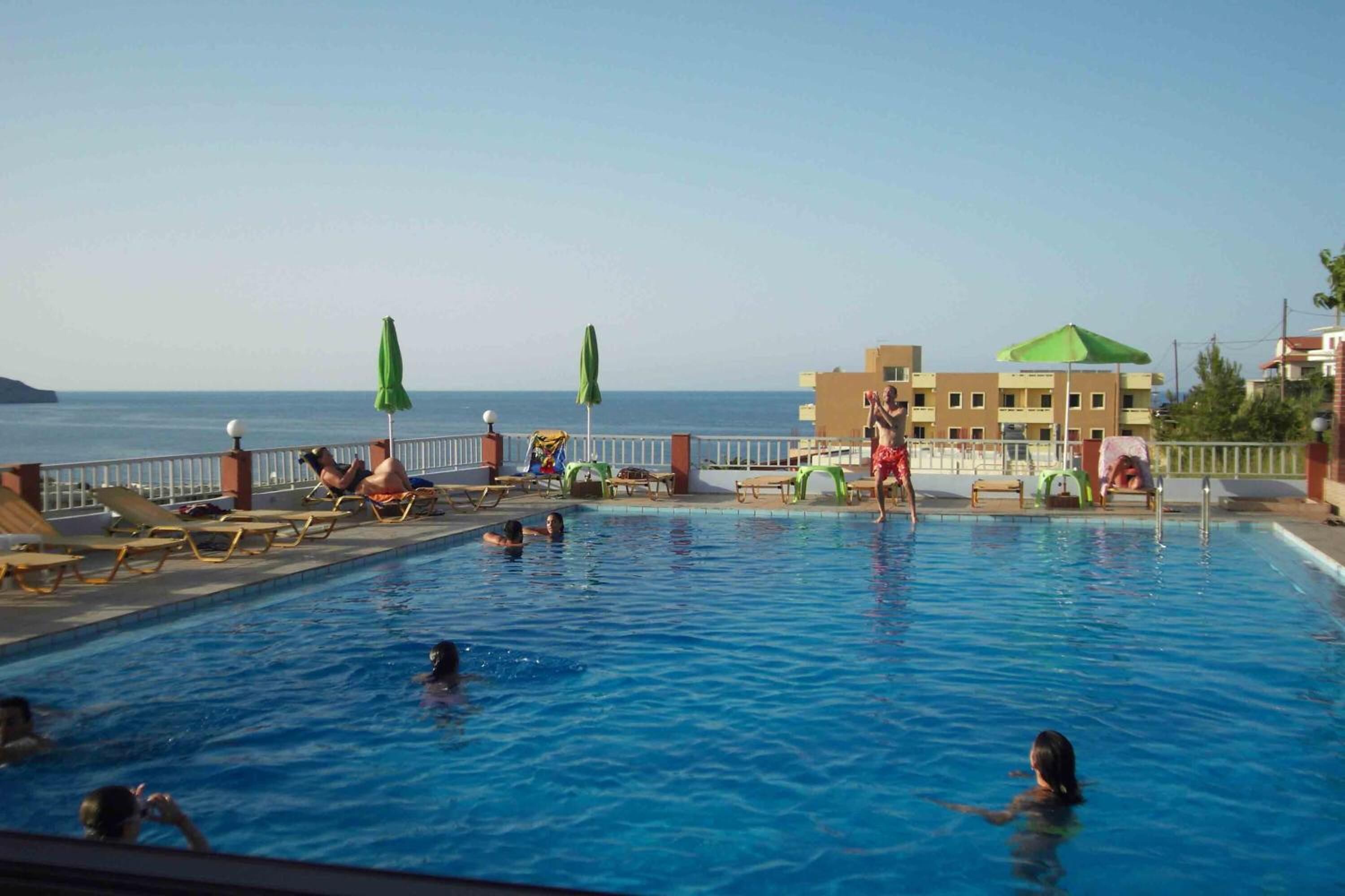 A swimming pool with lounge chairs and umbrellas, a building with a green roof, and a view of the sea.