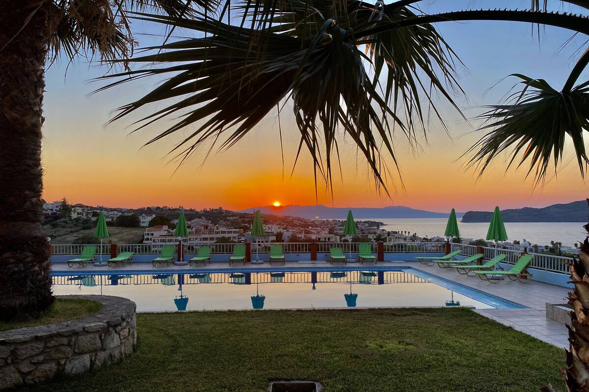 Zona junto a la piscina con sillones de descanso y sombrillas, con vista a una puesta de sol y una extensión de agua.