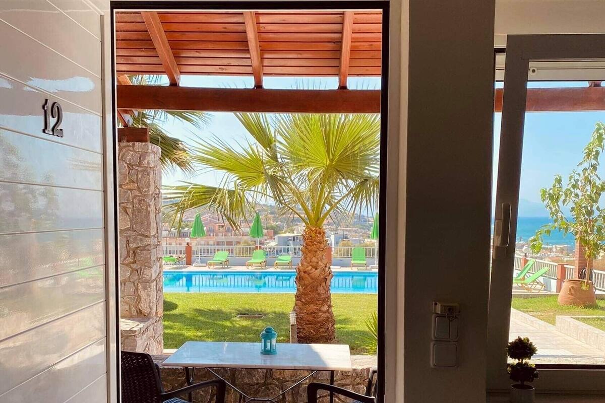 Puerta abierta que da a un patio con mesa y sillas, una palmera y vistas a una piscina y edificios.