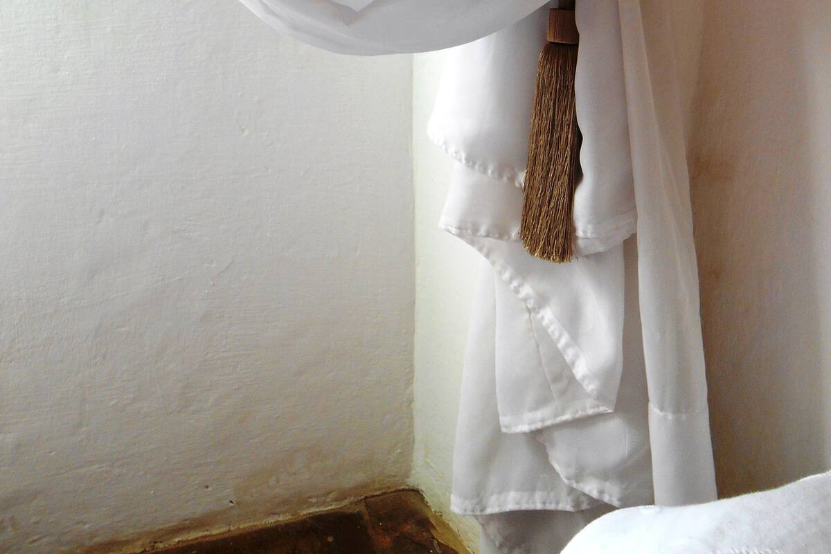A room corner with white walls, a window with white curtains tied back, and a wooden stick hanging on the wall.