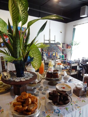 A dining table with various food items, a large potted plant, and a painting in the background.