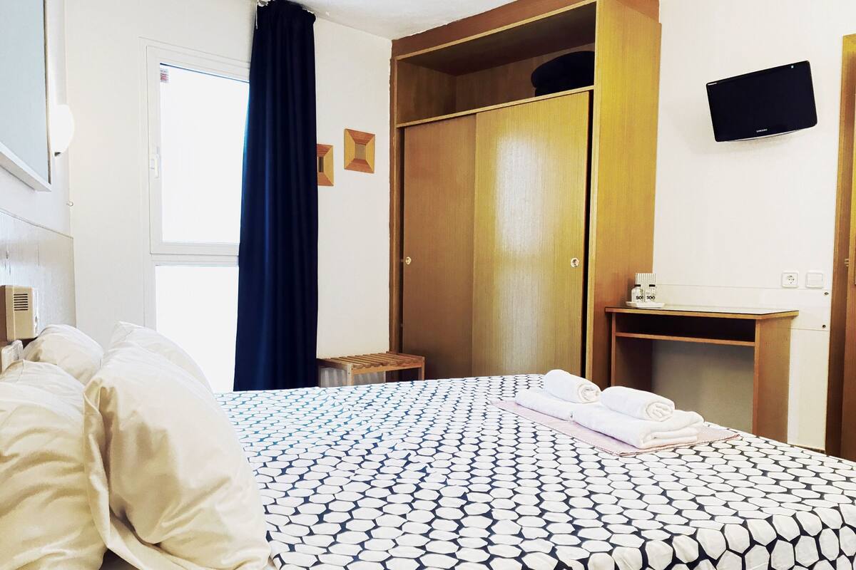 Habitació d'hotel amb un llit, un armari de fusta, una taula petita amb productes de tocador i un televisor fixat a la paret.