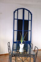 Una mesa con cubierta de vidrio y una planta en maceta, rodeada de sillas, frente a una pared con un ventanal de marco azul.