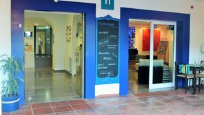 Entrada de un hotel con una decoración azul y blanca, un cartel que enumera servicios como pastanagas, hamburguesas y batidos, y un piso de baldosas.