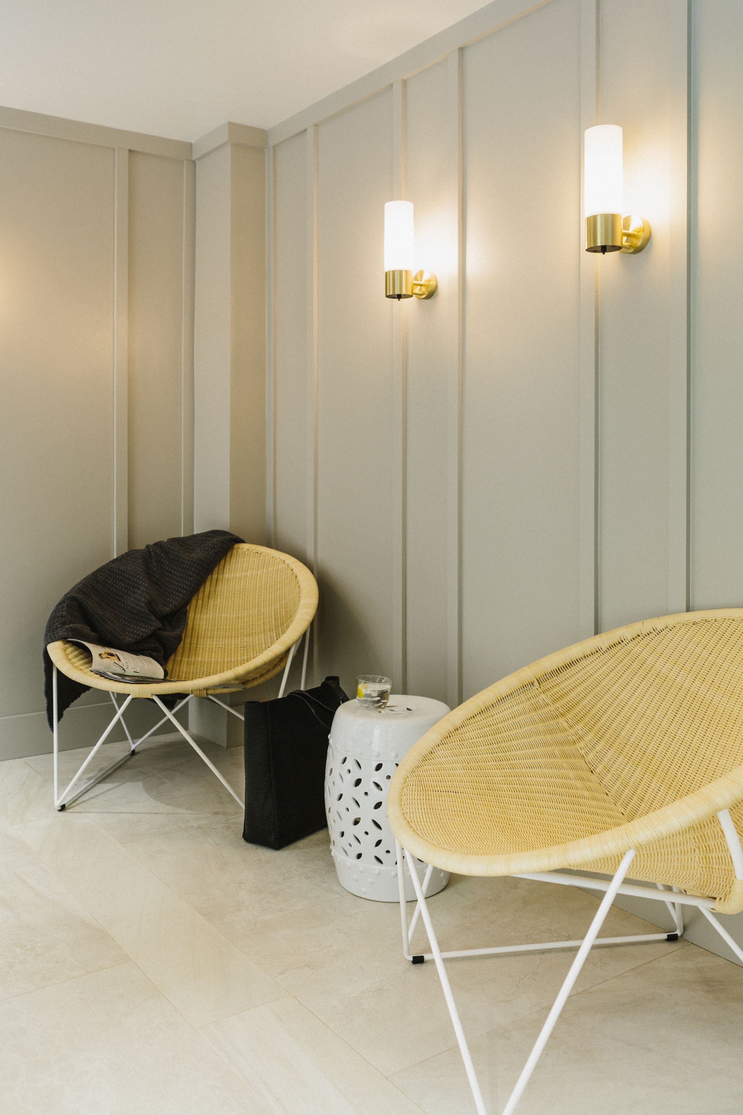 A room with two wicker chairs, one with a blanket, a small table with a glass, and wall-mounted lights.