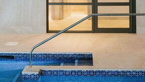 A modern spa area with a glass door leading to a treatment room, a sunburst light fixture on the wall, and a pool with blue-tiled steps. - Merchants Manor (Falmouth)