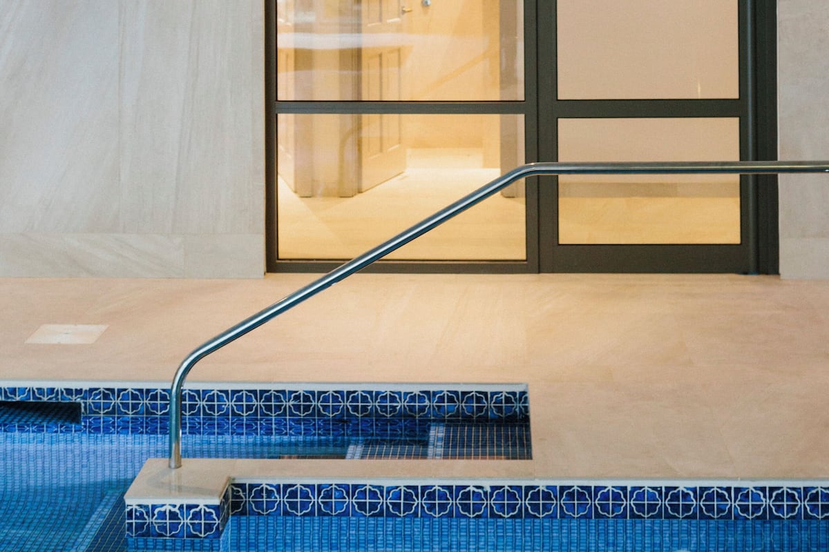 Un espace spa moderne doté d’une porte vitrée donnant accès à une salle de soins, d’un luminaire mural en forme de soleil et d’une piscine avec des marches recouvertes de carreaux bleus.