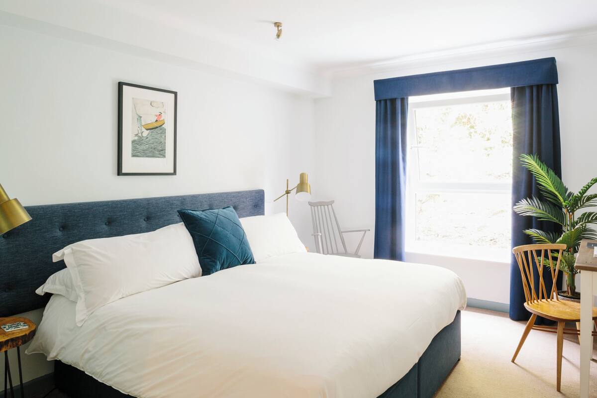 Un lit bien fait, avec une literie blanche et turquoise, un tableau encadré au mur, une chaise et une fenêtre avec des rideaux.
