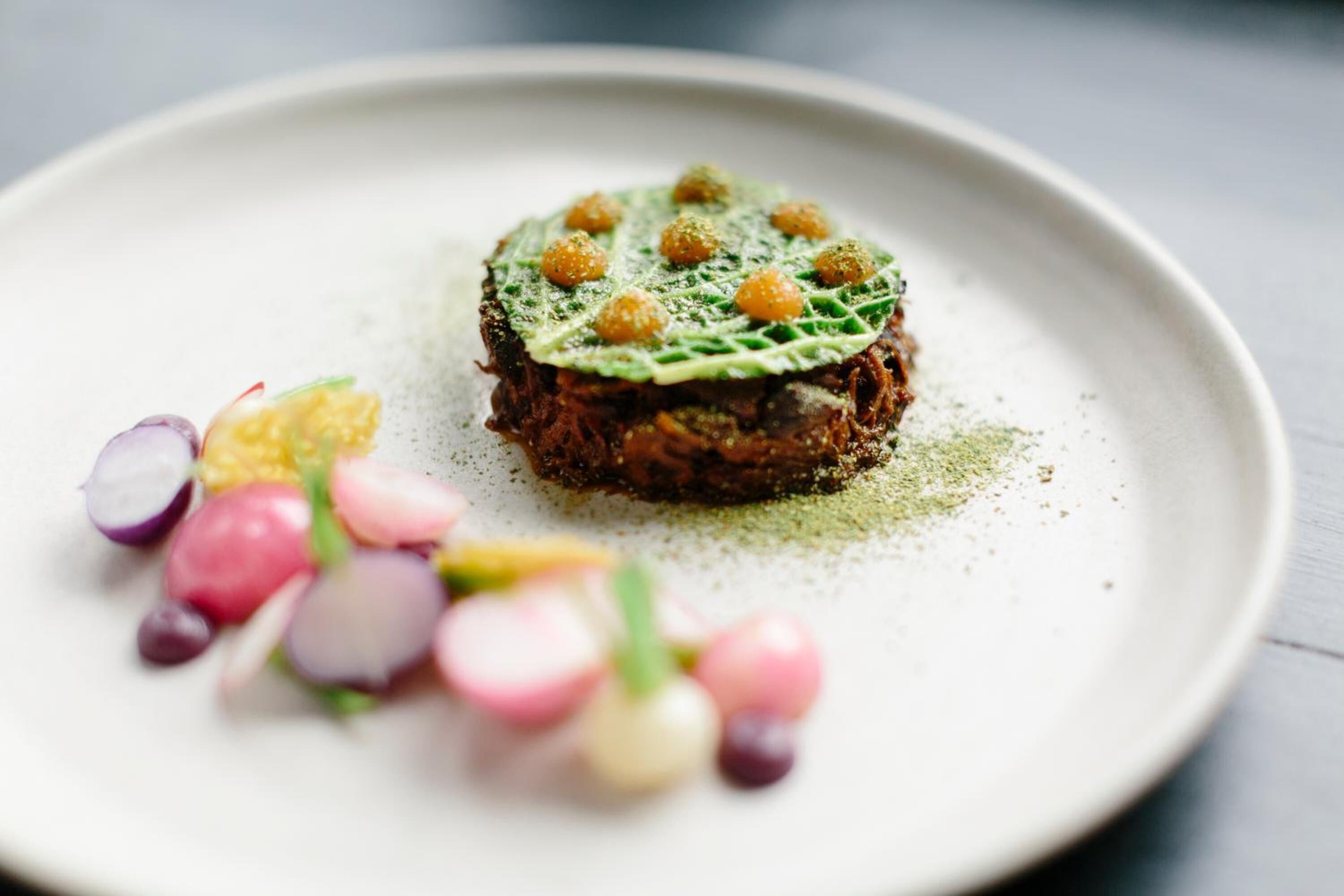 A dish with a central mound of food, surrounded by a green leaf and garnished with small round items, on a white plate.