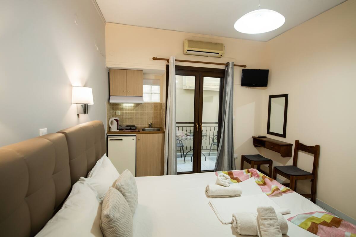 Quarto de hotel com cama, uma kitchenette pequena, varanda com mesa e cadeiras, e televisão fixada na parede.