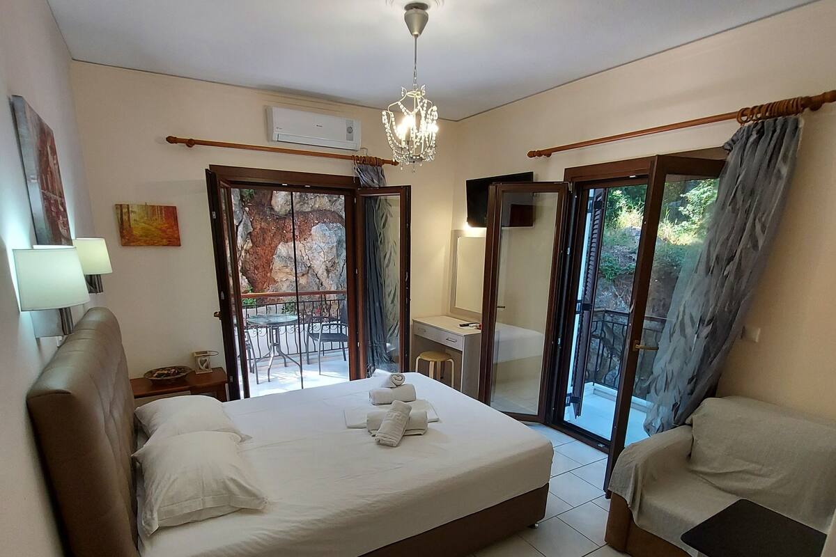 Quarto de hotel com cama, escrivaninha, cadeira, mesa pequena, lustre e varanda com vista para o exterior.