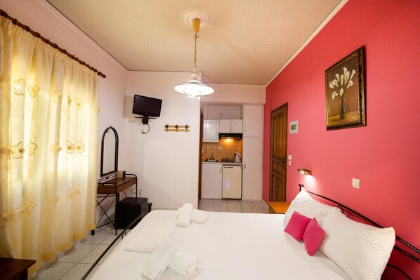 A hotel room with a bed, a desk, a mirror, a TV, and a kitchenette in the background.