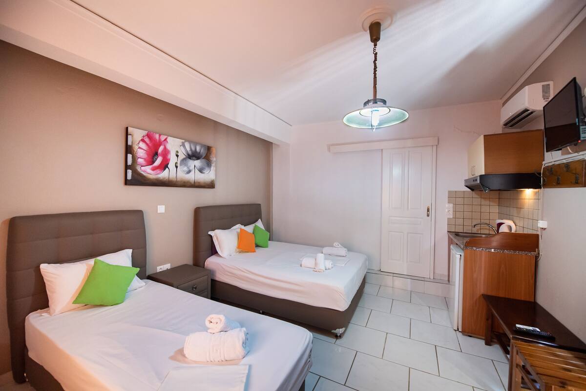 Quarto de hotel com duas camas, uma cozinha compacta, ventoinha no teto e um quadro na parede.