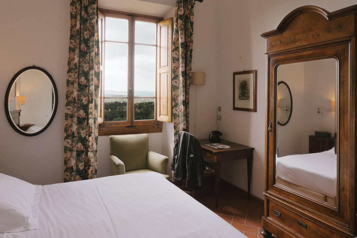 A hotel room with a bed, a desk, a chair, a large mirror, and a window with curtains.