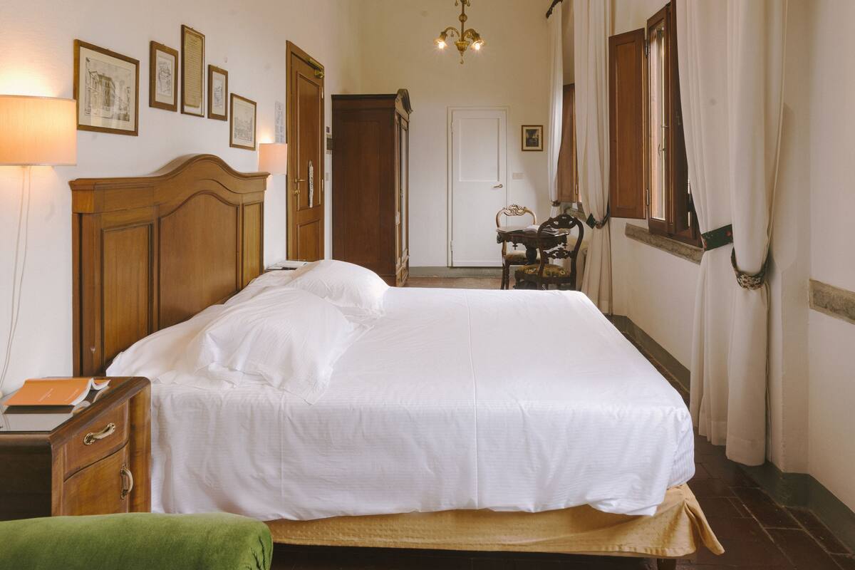 A neatly made bed in a room with a wooden headboard, a nightstand, a green chair, and a desk with a lamp.