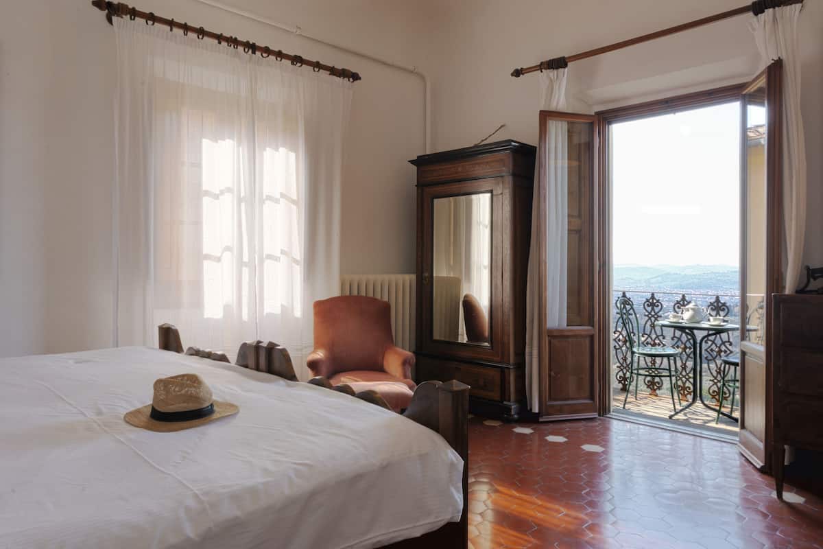 A hotel room with a large bed, a wooden chair, a mirror, and a balcony with a view.