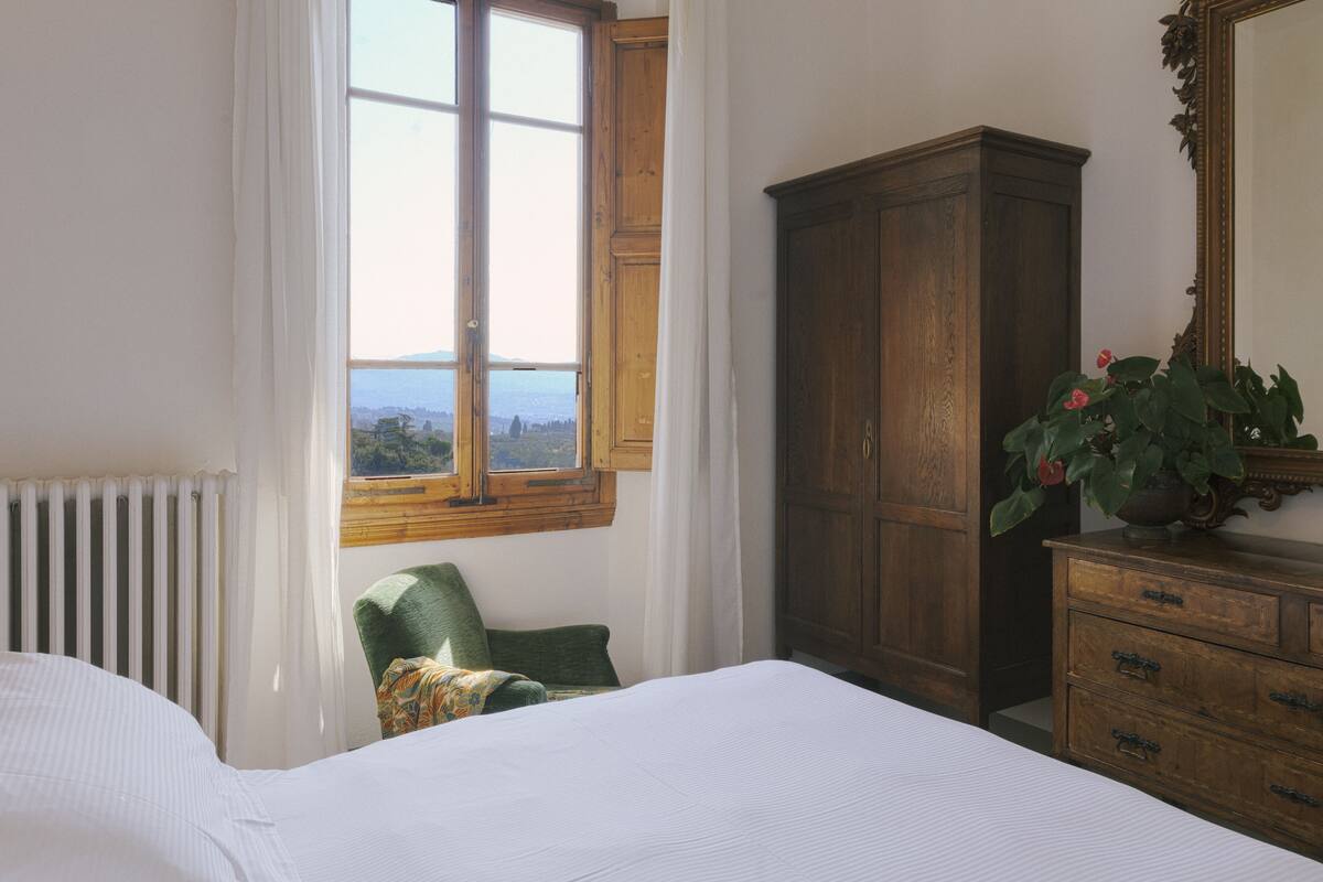 A bedroom with a large bed, a wooden wardrobe, a green chair, and a window with a view of the mountains.