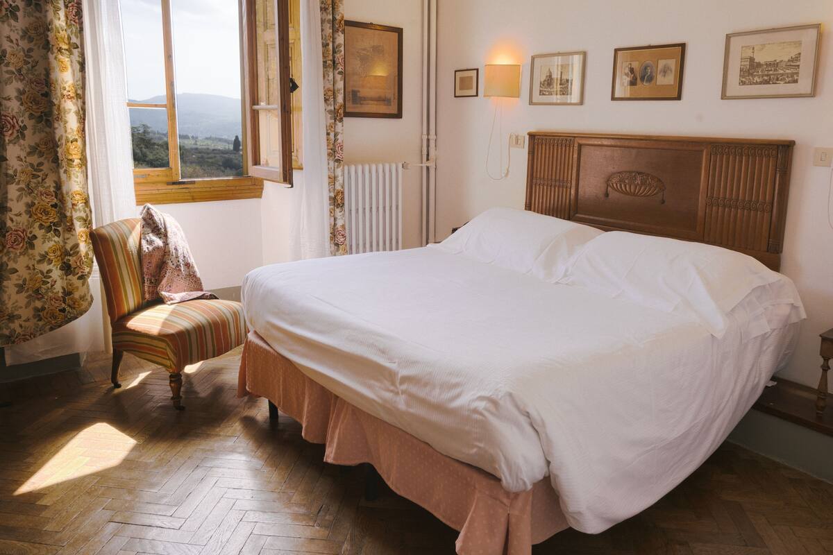 A bedroom with a large bed, a chair, a window with a view, and framed pictures on the wall.