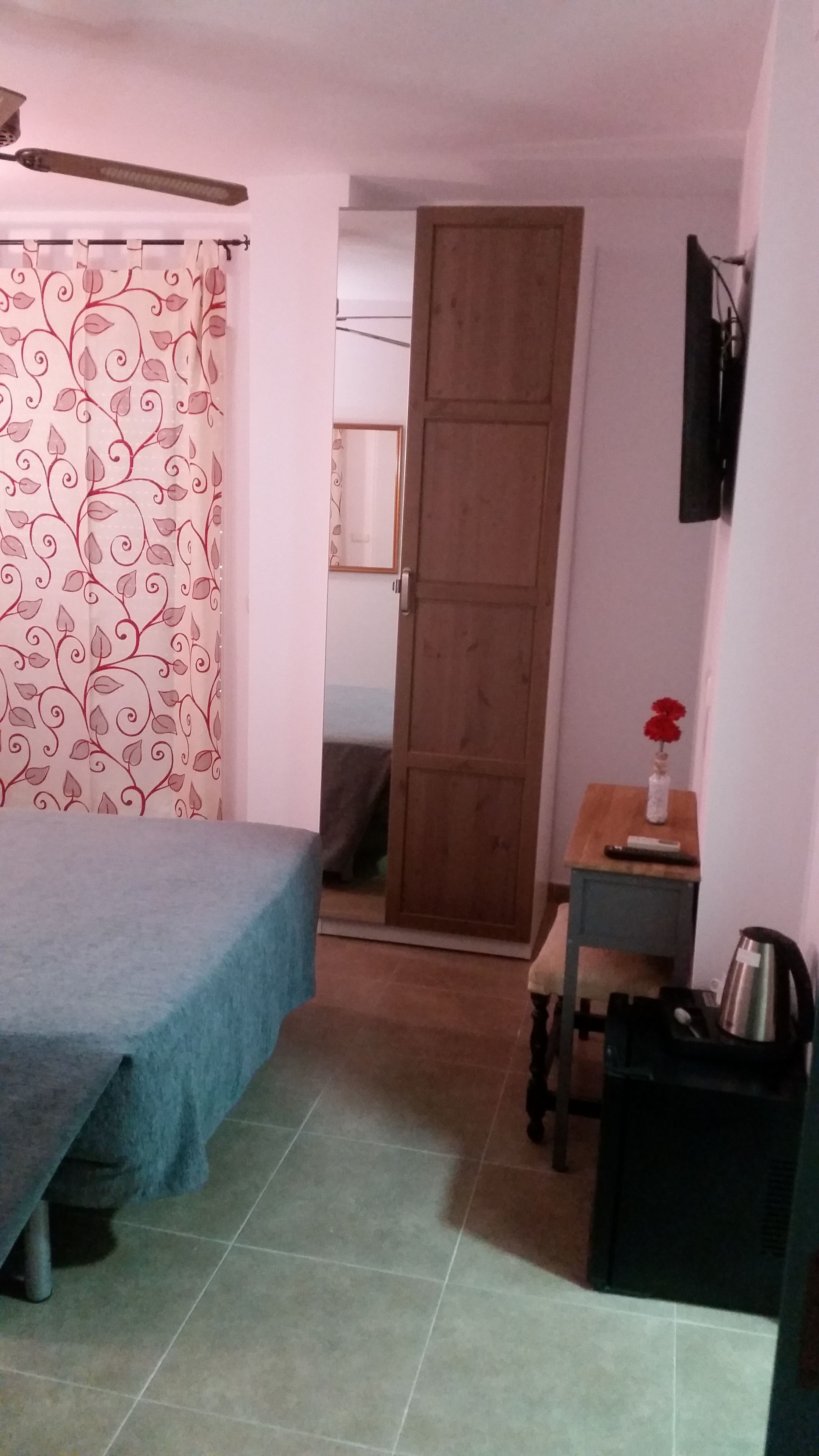 Una stanza con un letto, una porta in legno, un tavolino con un vaso di fiori e un bollitore su un ripiano.