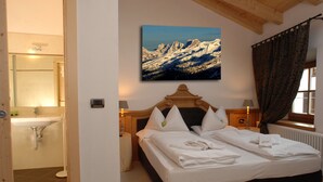A bedroom with a bed, a wooden headboard, a painting of mountains, a window with curtains, and a bathroom with a sink and mirror. - Excelsior (Cavalese)