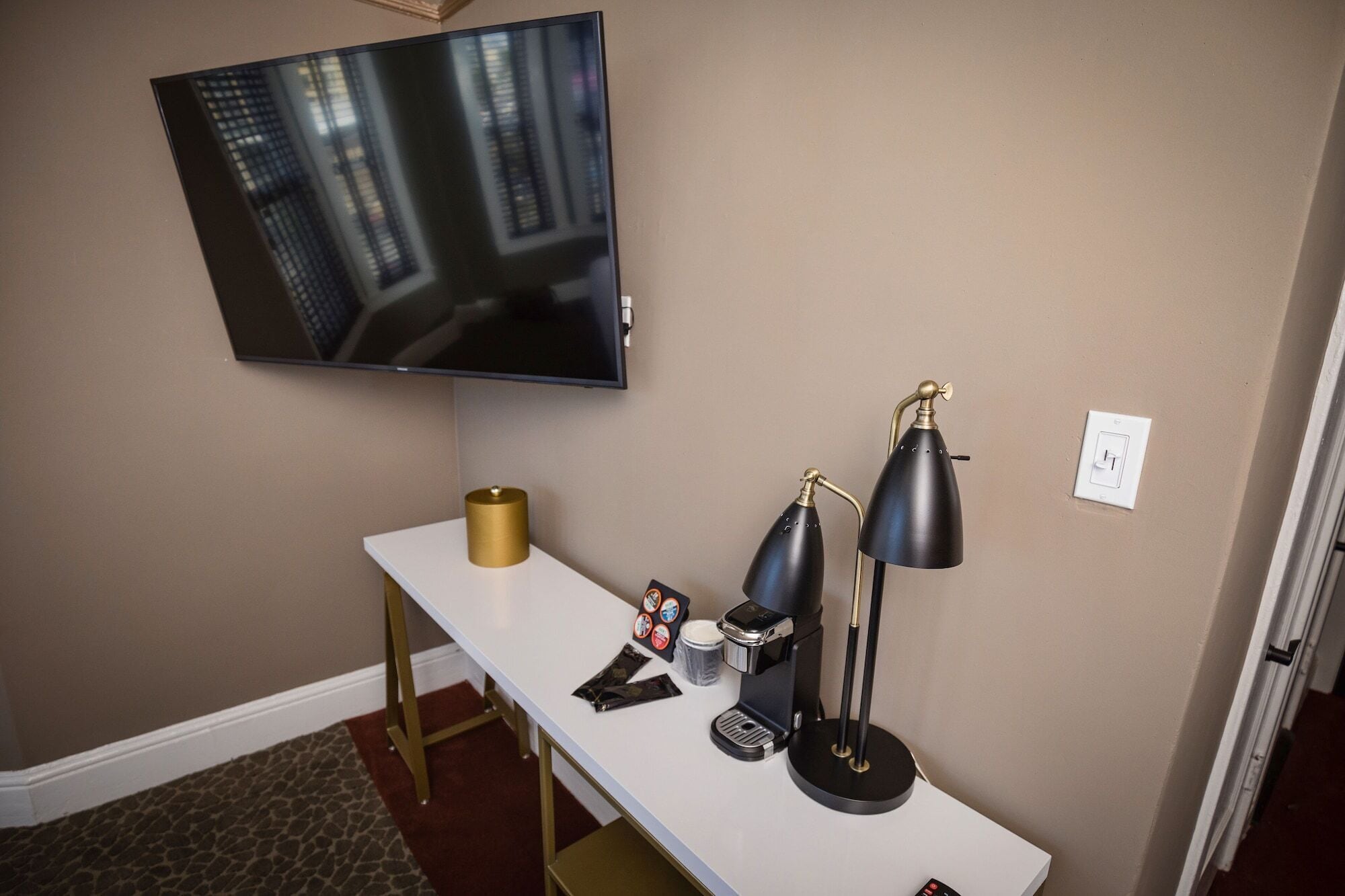 A hotel room with a flat-screen TV mounted on the wall, a white desk with a coffee maker, two black desk lamps, and a gold-colored cylindrical object on the desk.
