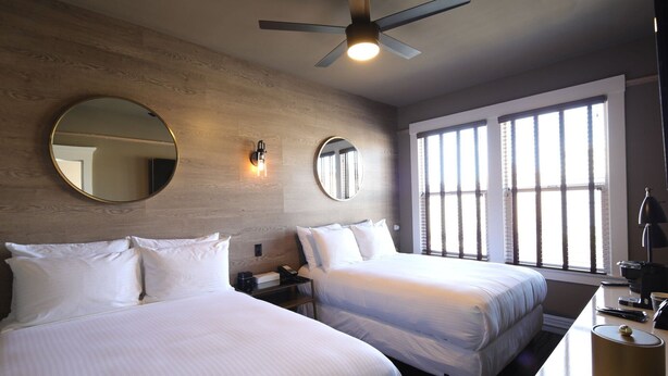 Habitación de hotel con dos camas, un escritorio, un ventilador de techo y ventanas amplias.
