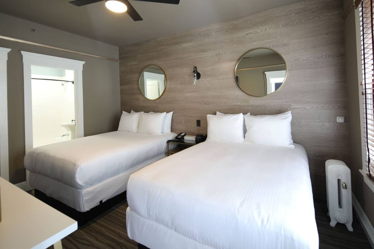 Une chambre d’hôtel avec deux lits, un bureau, une chaise, un miroir et un ventilateur de plafond.