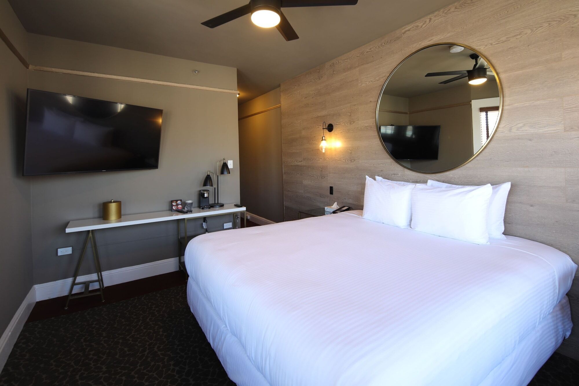 A hotel room with a large bed, a desk with a lamp, a flat-screen TV, and a ceiling fan.
