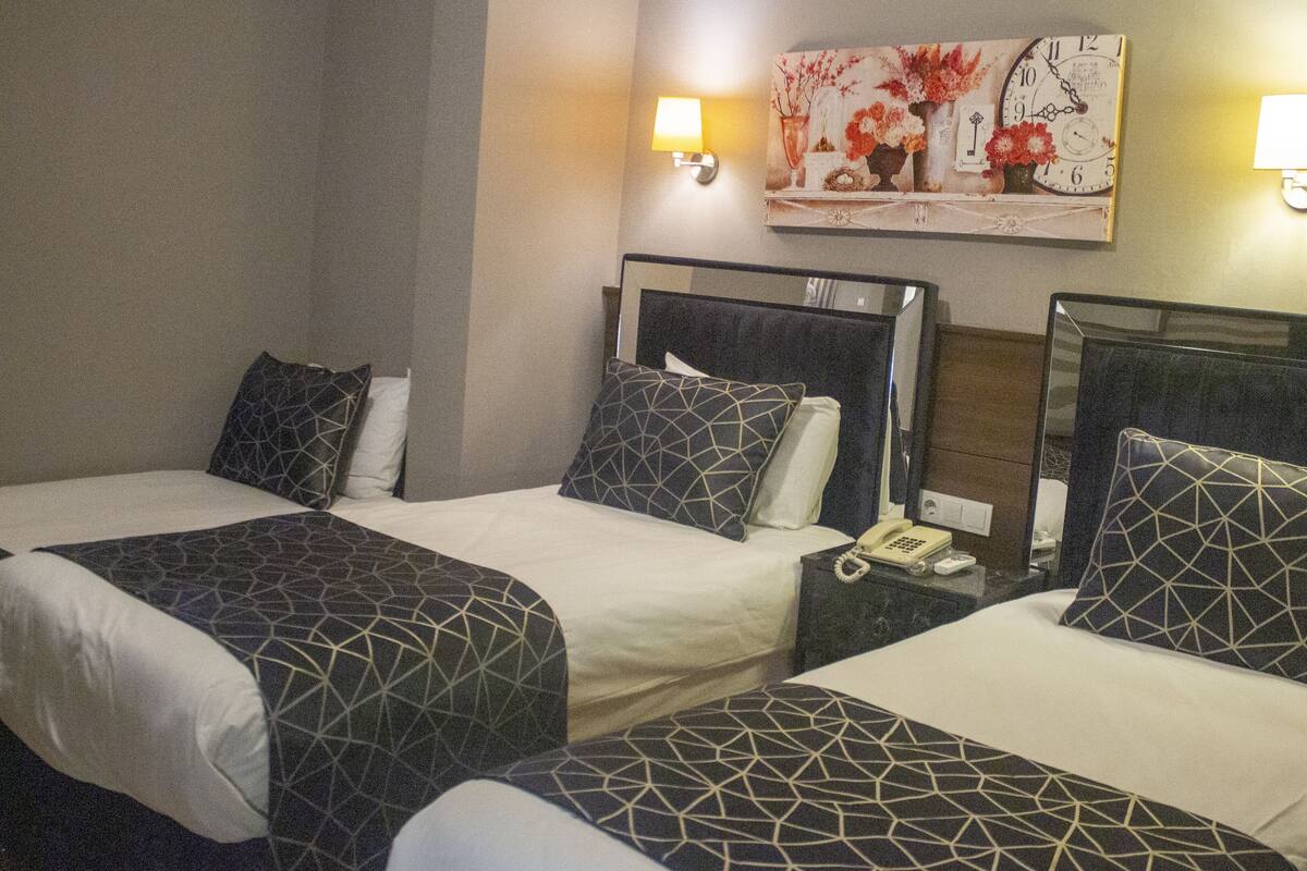 A hotel room with two beds, each with a geometric-patterned bedspread and matching pillows. There are bedside tables with lamps and a wall-mounted clock.