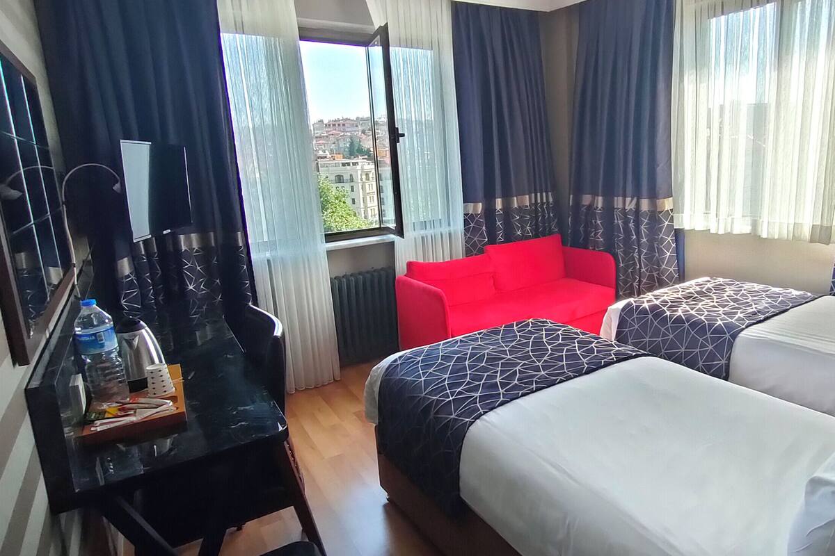A hotel room with a bed, a red chair, a desk with a water bottle, and a view of the city through the window.
