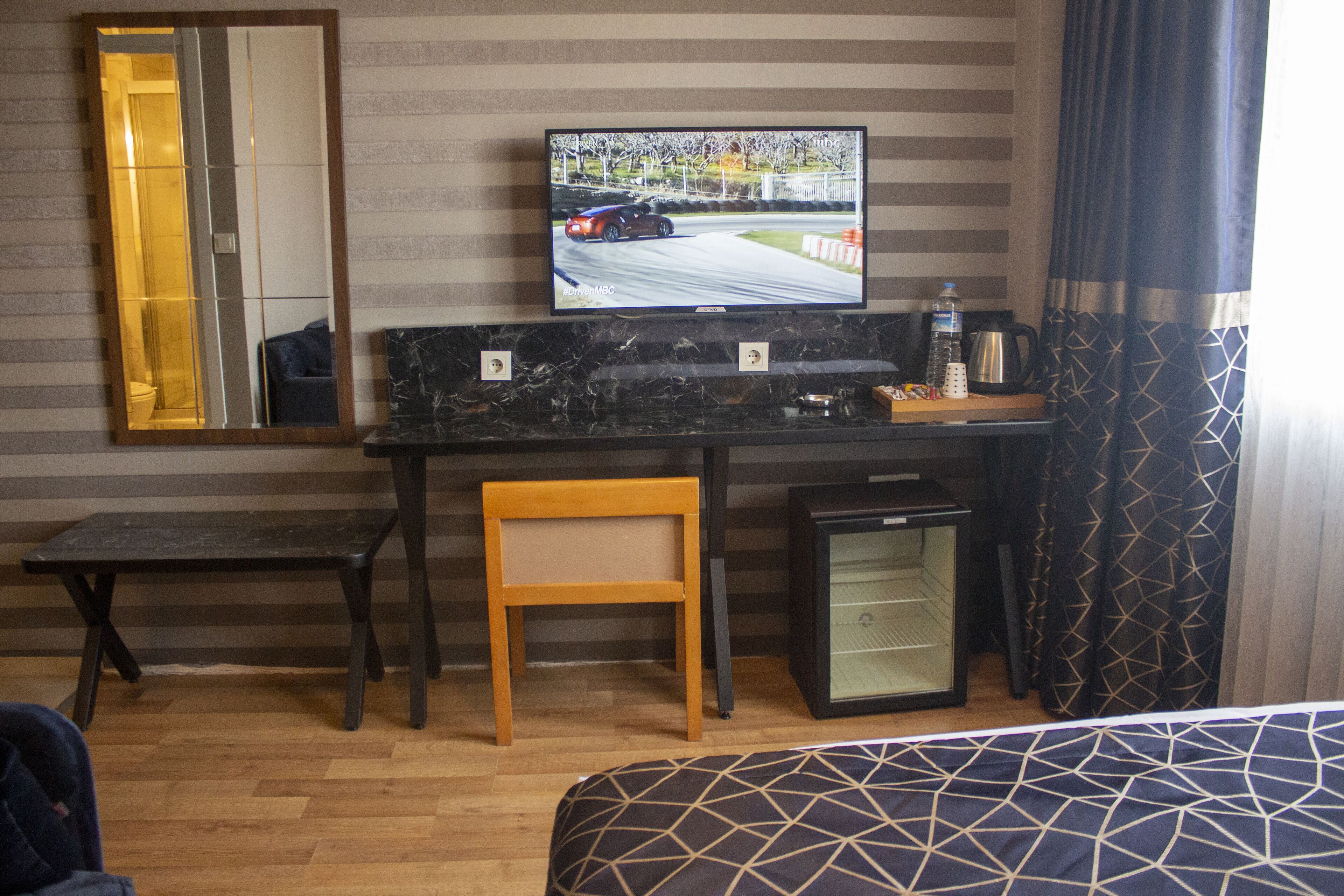 A hotel room with a bed, a desk with a television, a small refrigerator, and a mirror on the wall.