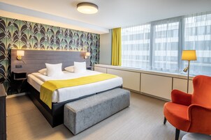 A hotel room with a bed, bedside tables, a bench, a chair, a window with curtains, and a wall with a leafy pattern.