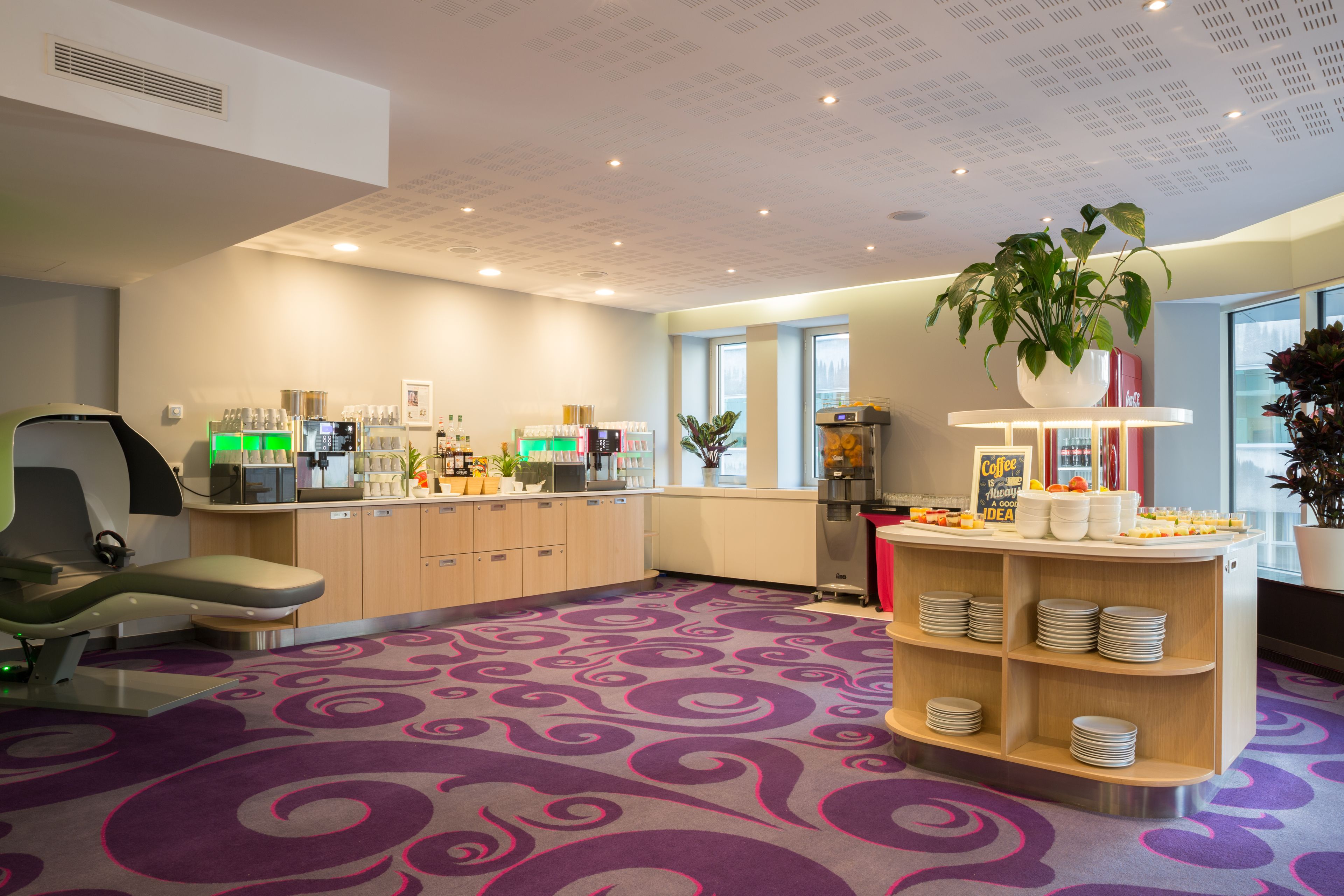 A modern hotel lobby with a purple carpet, a reception desk, a coffee station, and a plant.