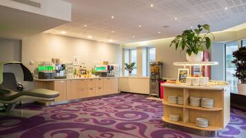 A modern hotel lobby with a purple carpet, a reception desk, a coffee station, and a plant.