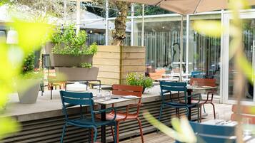 Un área de comedor al aire libre con varias mesas y sillas, rodeada de plantas en macetas y una estructura de madera.