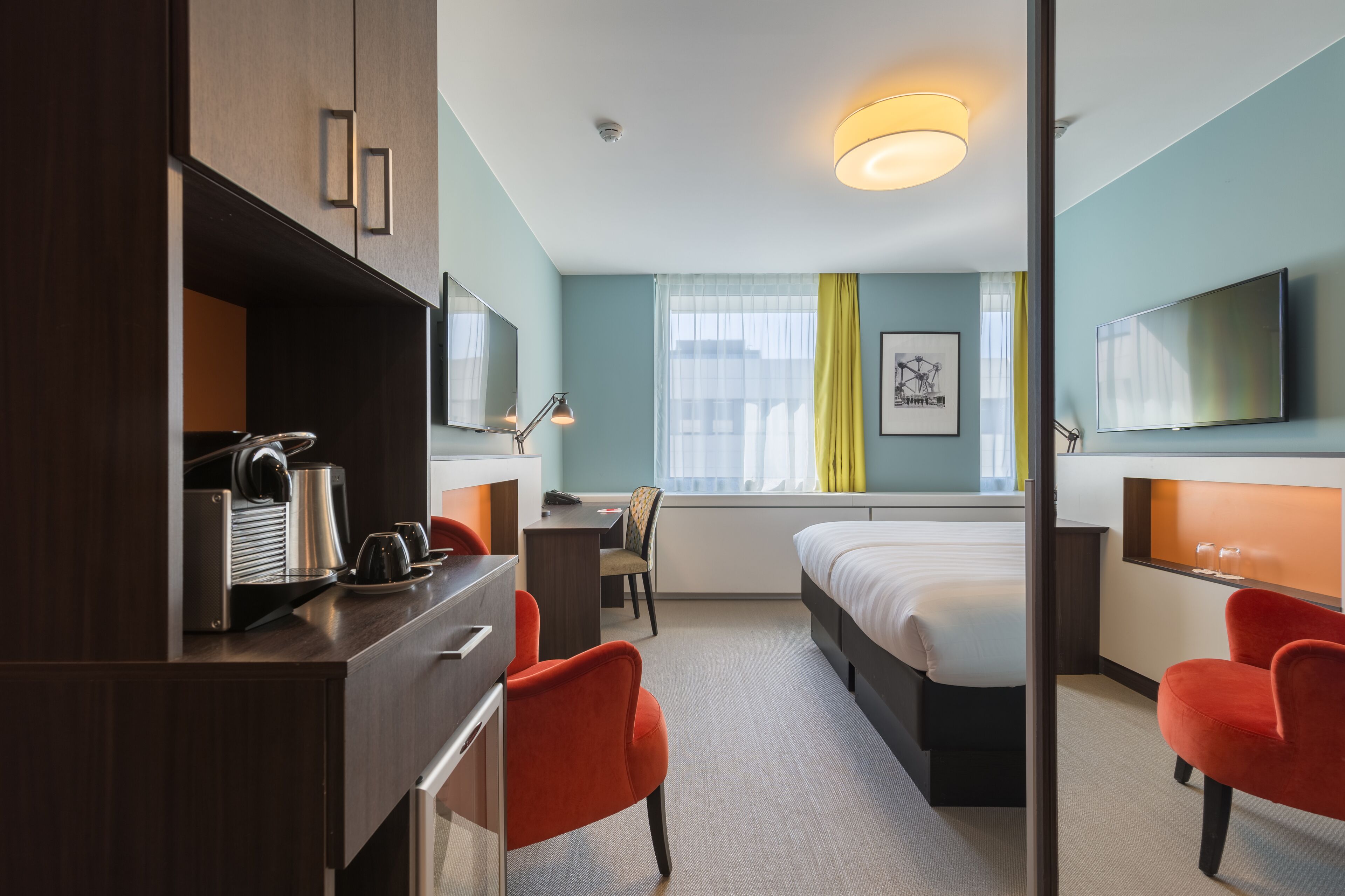 A hotel room with a bed, a desk with a coffee maker, two red chairs, a TV, and a window with curtains.