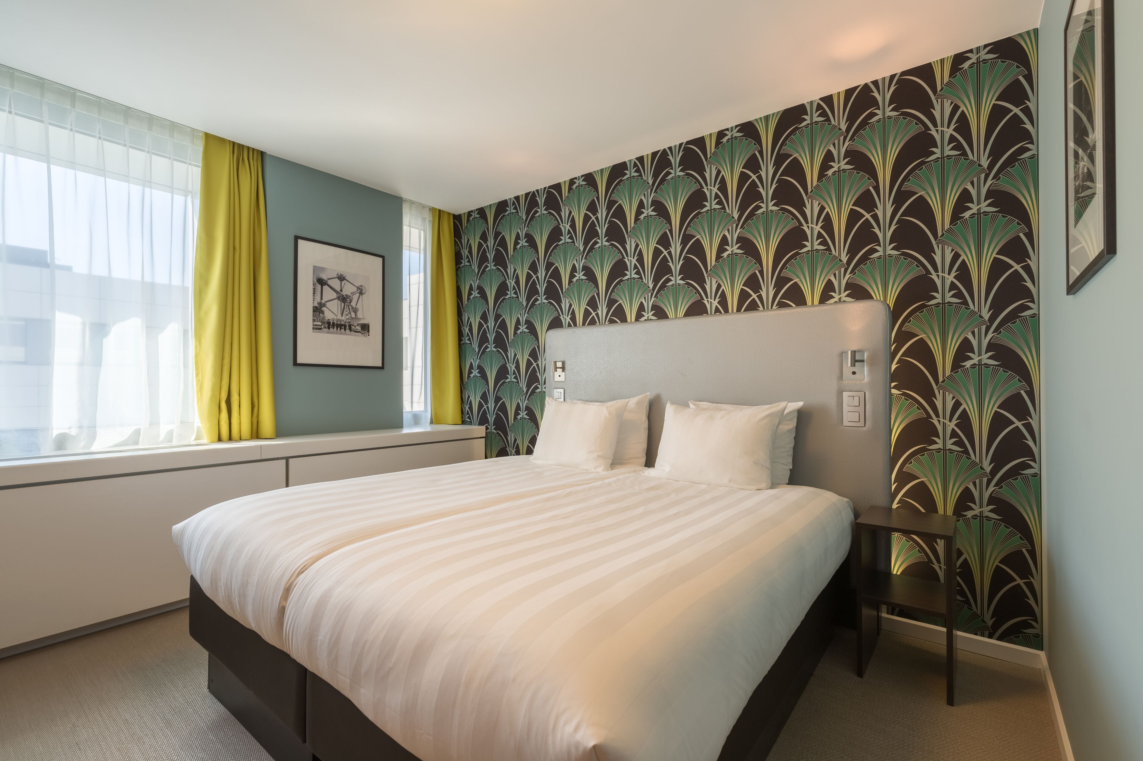 A neatly made bed in a hotel room with a large window, a framed picture on the wall, and a patterned wallpaper.
