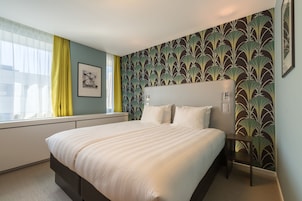 Una cama bien tendida en una habitación de hotel con un ventanal grande, un cuadro en la pared y un papel tapiz estampado.